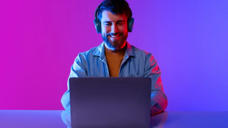 Happy Millennial Guy Wearing Headphones Browsing Internet On Laptop, Studio